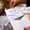 Un miembro de la mesa electoral muestra un voto nulo con el dibujo de una paloma y el lema "paz" en varios idiomas